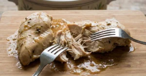 Tender cooked chicken being shredded on a cutting board using two forks. Juicy texture visible, strands pulling apart easily. Crockpot with flavorful broth in the background.