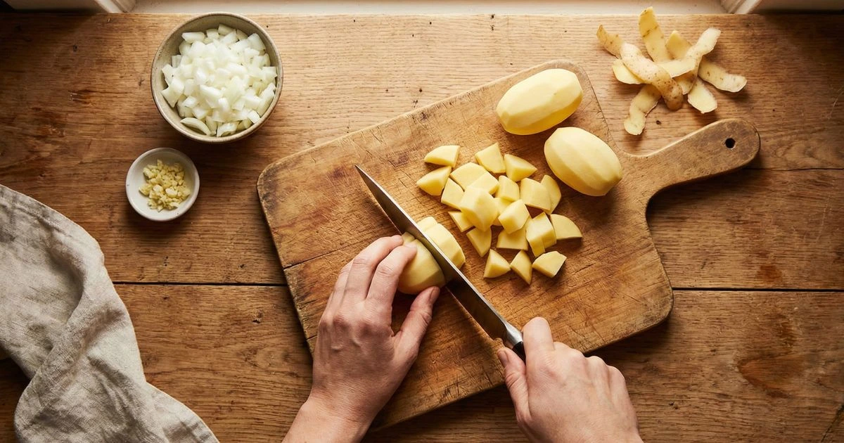 Overhead kitchen scene with peeled potatoes being diced on a wooden cutting board, chopped white onions in a bowl, minced garlic in a small dish, chef’s knife in hand, warm natural lighting, rustic home kitchen style