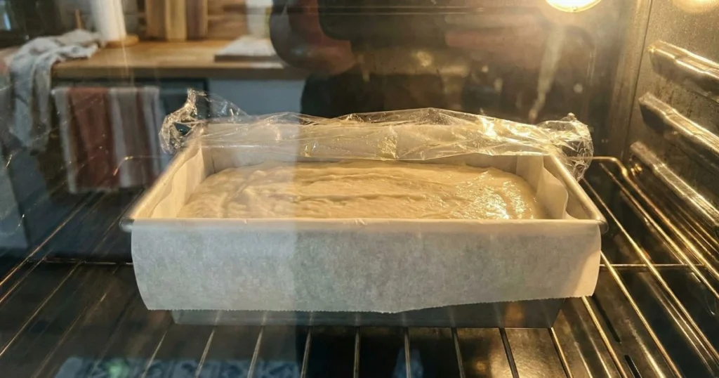 Gluten-free bread batter spread in a parchment-lined loaf pan, smoothed top with slight sheen of moisture, loosely covered with oiled plastic wrap, placed in a warm oven with light on.